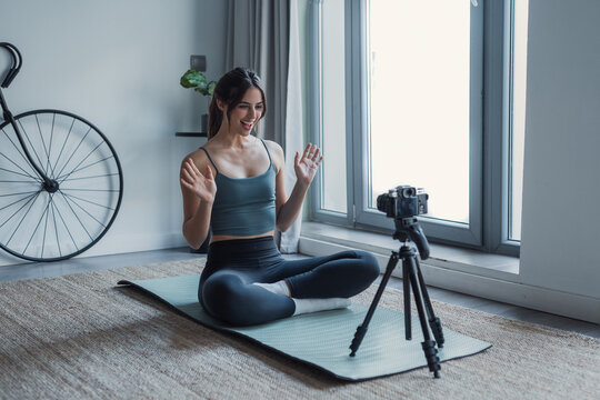 A smiling young woman sits cross-legged on a yoga mat by a bright window, leading a home fitness and yoga session while recording a vlog on a tripod camera, expressing wellness, motivation and calm.