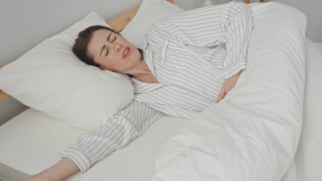 Woman clutching abdomen with hand for stomachache while lying on bed in bedroom, striped pajamas and white duvet visible; morning pain.