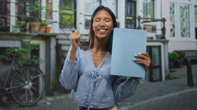 Thai woman holds blue clipboard and points pen to it while smiling with a thumbs up on a cobblestone street; confidence.