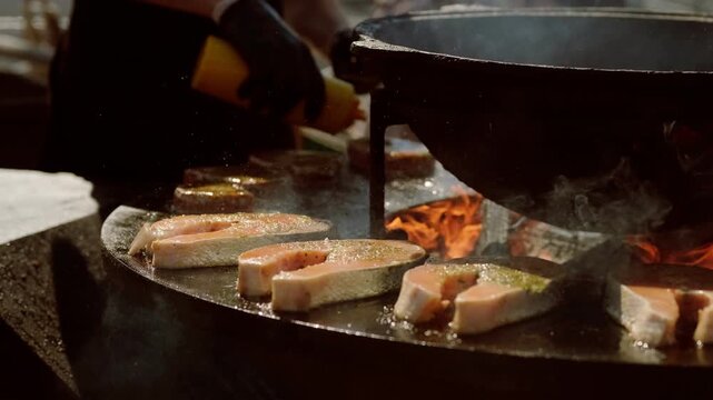 Chef cooking salmon steak on outdoor grill with open flames. Professional grilling process of fish fillet. Culinary event, street food preparation and gourmet seafood cooking technique.