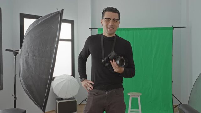 Man holding camera and wearing glasses in studio, smiling while posing by green screen with softbox and stool visible; confidence creativity.