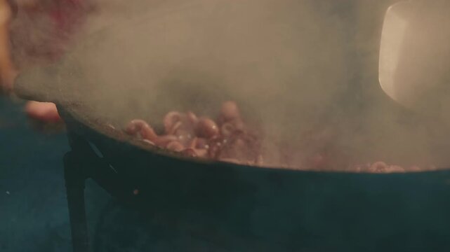 Chef cooking octopus in large cast iron wok over fire. Street food preparation sequence showing steaming seafood dish being stirred in outdoor commercial kitchen.