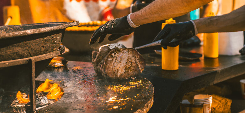 Chef grilling large piece of pork meat on open fire barbecue. Man cooking seasoned steak with metal utensil outdoors. Concept of street food and culinary skill.