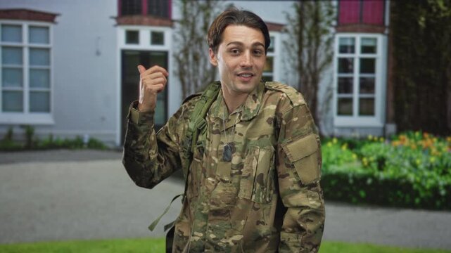 Man in camouflage uniform wearing dogtags, bare thumb pointing over shoulder in a casual salute gesture toward a building entrance, smiling; pride duty.