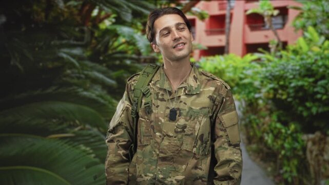 Man in military camouflage with dog tags and shoulder strap smiling, head tilted, standing on a lush garden path beside a red building; duty pride.