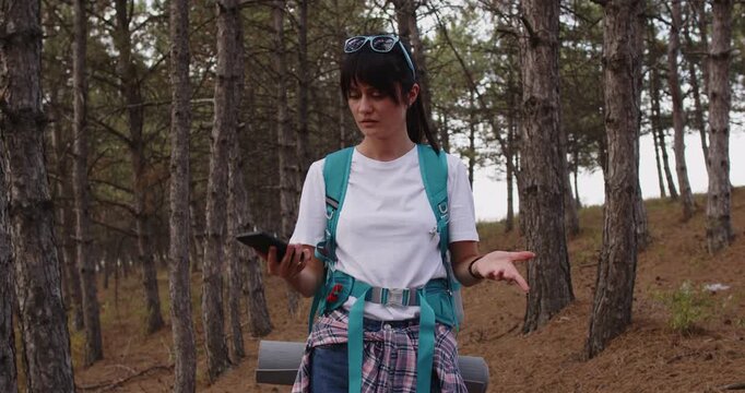 Tourist backpacker checks smartphone map, lost in forest. With no signal, navigation and geolocation fail, leaving her unsure of route and coverage. Theme offline travel and adventure.