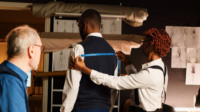 Tailors team taking measurements for client, working in atelier to create custom made tailored suit. Couturier and assistant measuring customer to design perfect fit clothes. Handheld shot.