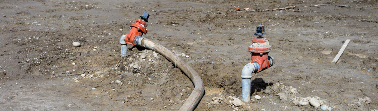 Construction site at groundworks stage, raw dirt with water utility pipes, shutoff valves, gate valve backflow preventer, connected to temporary industrial hose pumping out water
