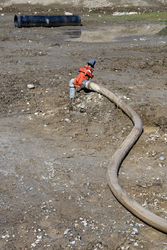 Construction site at groundworks stage, raw dirt with water utility pipes, shutoff valves, gate valve backflow preventer, connected to temporary industrial hose pumping out water
