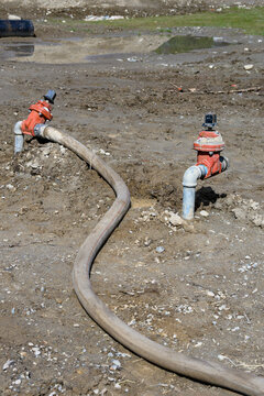 Construction site at groundworks stage, raw dirt with water utility pipes, shutoff valves, gate valve backflow preventer, connected to temporary industrial hose pumping out water
