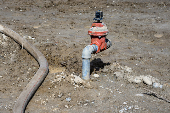 Construction site at groundworks stage, raw dirt with water utility pipes, shutoff valves, gate valve backflow preventer, connected to temporary industrial hose pumping out water
