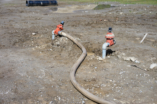 Construction site at groundworks stage, raw dirt with water utility pipes, shutoff valves, gate valve backflow preventer, connected to temporary industrial hose pumping out water
