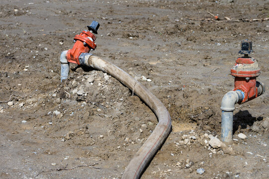 Construction site at groundworks stage, raw dirt with water utility pipes, shutoff valves, gate valve backflow preventer, connected to temporary industrial hose pumping out water
