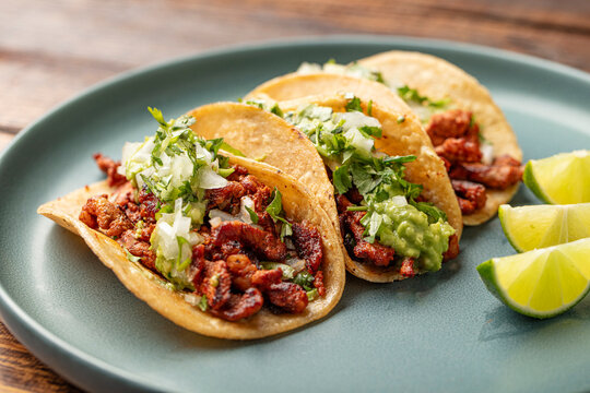 Close up shot of three grilled pork tacos with corn tortillas, onions, guacamole and cilantro on a green plate and wooden table