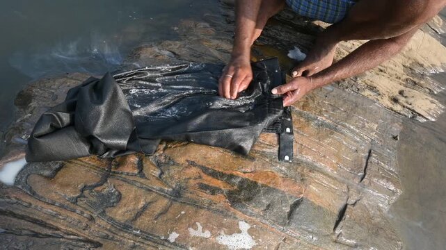 A man is washing clothes by hand on a stone at the riverbank. He uses soap to rub the fabric against the surface of the stone. How to wash clothes in the traditional way.