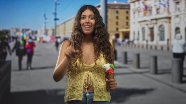 Woman in yellow crochet top smiling, holding a triple scoop ice cream cone and pointing finger to camera on a sunny urban street with historic building and bollards; summer joy.