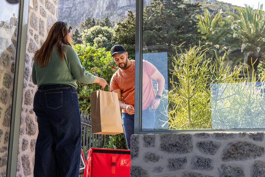 Courier and customer exchanging paper bag at porch, courier in orange cap with red insulated bag