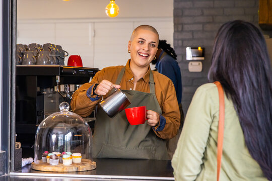 Barista in olive green apron pouring milk from metal pitcher into red mug, customer holding bag