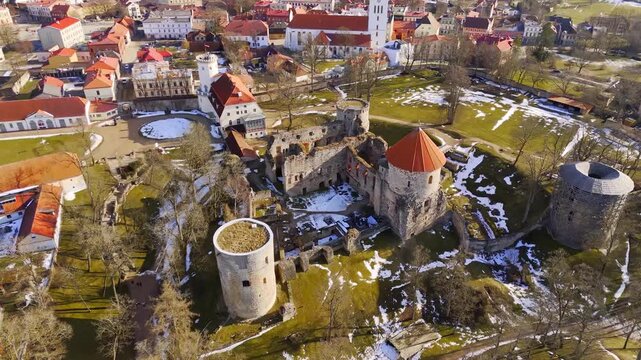 Aerial view of Cesis Castle ruins with round towers and St. John&rsquo;s Church in the background, showing layered heritage, snow patches, and historic town fabric in spring light.