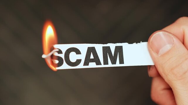Burning &ldquo;scam&rdquo; label held by hand, symbolizing scam awareness, fraud prevention, consumer protection, and rejecting deceptive schemes or financial traps.