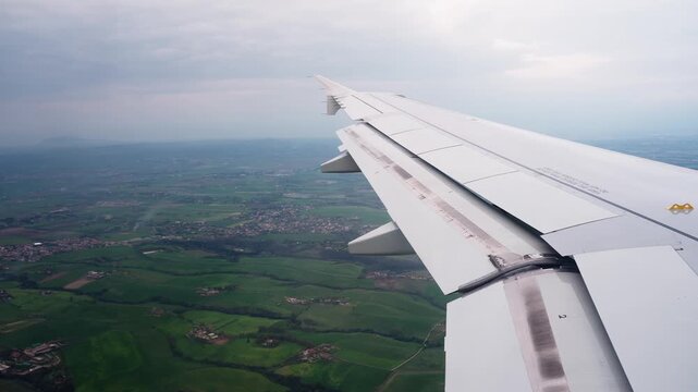 Airplane wing view above green countryside during descent, showing a passenger perspective of commercial air travel over patchwork fields beneath a cloudy sky.