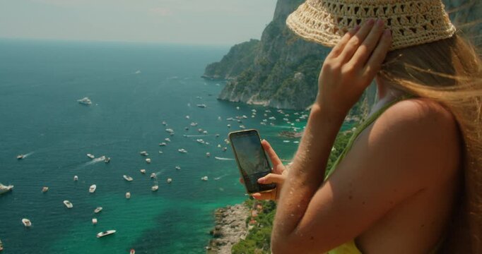 Woman taking a phone photo overlooking Marina Piccola cliffs and boats in summer Capri Italy