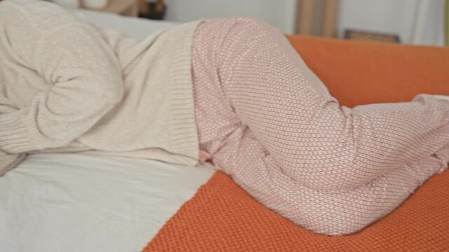 Woman lying on bed in bedroom wearing sweater and patterned pajamas, hand near face with a soft smile and relaxed eyes; restful serenity.