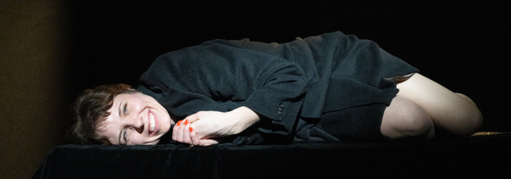 In this image, a woman lies on a dark stage, laughing joyously, embodying a carefree spirit, contrasting with earlier somberness and inviting positivity through her vibrant expression.
