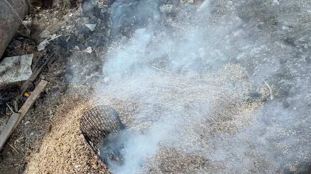 Traditional rice husk carbonization process in a pyramid pile with smoke coming out from the chimney.