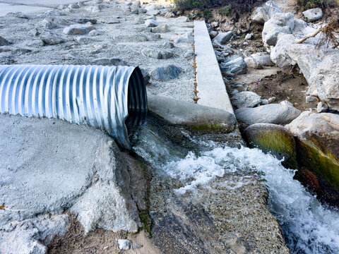 Water flows out of a corrugated metal pipe onto rocky concrete and stone surrounded by large boulders and sparse vegetation creating an energetic drainage scene outdoors
