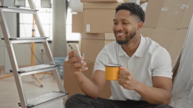 Man holding smartphone and yellow mug, smiling while seated among stacked moving boxes in an indoor building; contentment moving.