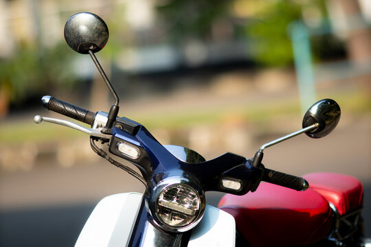 Modern scooter with sleek chrome handlebars and round mirrors. Red seat contrasts with white body and metallic accents. Circular headlight gleams under soft daylight.
