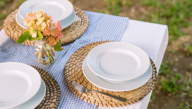 Elegant outdoor dining setup with rustic woven placemats, delicate white plates, and a small vase of flowers on a white tablecloth