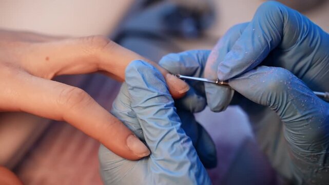 Macro salon shot of a gloved nail technician using a cuticle pusher to lift and clean the nail plate during detailed professional manicure preparation.