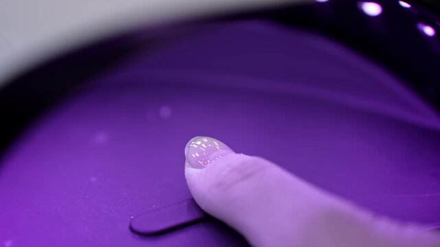 Glitter accent thumbnail cures under a UV LED nail lamp, capturing violet glow, gel hardening, and modern salon technology during a professional manicure.