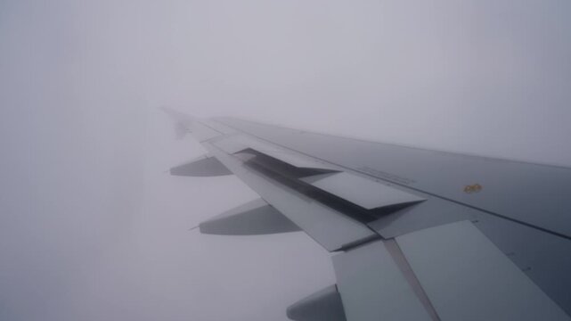 Airplane wing disappears into thick cloud during descent, capturing a whiteout flight moment with low visibility, muted light, and an eerie midair atmosphere.