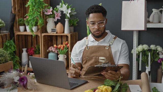 Man florist in apron writing on clipboard beside laptop in studio surrounded by vases and blooms; focused small business management.