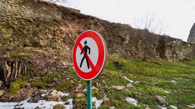No pedestrian access sign below a crumbling stone wall with loose rocks on a grassy slope, highlighting hazard, restricted area, and danger from falling masonry.