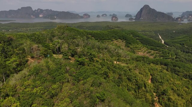 Stunning aerial perspective flying backward over a dense tropical forest and hills, slowly revealing the magnificent panorama of limestone karsts in thailand's iconic phang nga bay on a sunny day