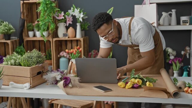 Man florist leaning over laptop typing at flower studio workbench surrounded by tulips, wooden crates and kraft wrapping paper; focused entrepreneurship.