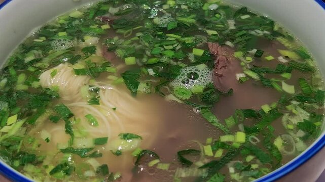 close-up steaming beef noodle soup with thin noodles, sliced beef, and chopped scallions in broth