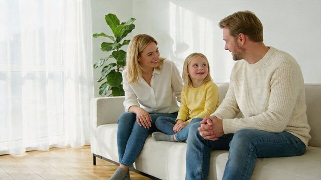 Happy Caucasian family mother father and daughter sitting on white sofa in living room bright home interior parent and child interaction conversation at home casual clothing