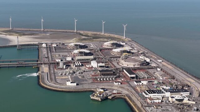 High angle perspective showing a liquefied natural gas import terminal and regasification plant with large storage tanks, situated next to a wind farm on a coastal industrial harbor peninsula