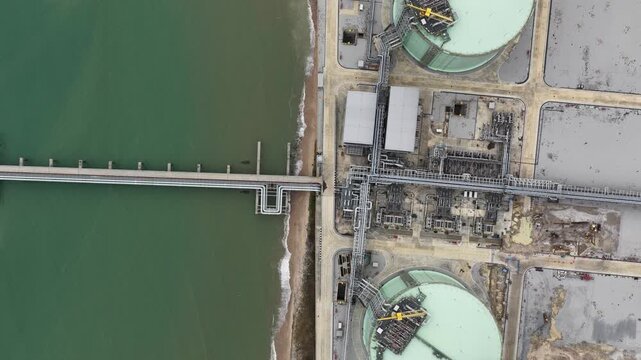 Aerial zenithal perspective showing the industrial infrastructure of a coastal lng plant, including large storage tanks, intricate piping, and a pier extending into the green hued sea