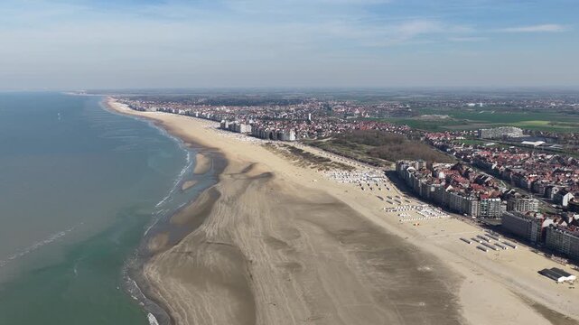 Incredible aerial perspective flying along a vast, sandy shoreline with gentle waves from the north sea lapping at the coast, showcasing the sprawling urban landscape of Knokke, Belgium, seaside town