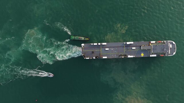 Aerial top down drone video of a tug boat escorting a large ship out of port, showing harbor operations, maritime transport, vessel guidance, and commercial shipping movement.