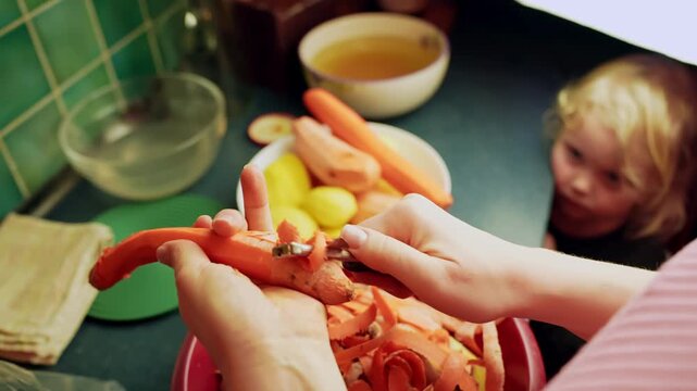 Hands peel a carrot with a peeler over a bowl on kitchen counter while a small child watches nearby. Cozy family meal preparation at home.
