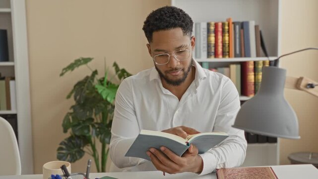 Man reading a book with finger tracing a page at a desk by a bookshelf and lamp in a building; quiet concentration study.