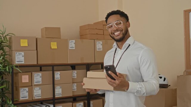 Man holding parcel and smartphone in hand, smiling and presenting packages in building; friendly service.