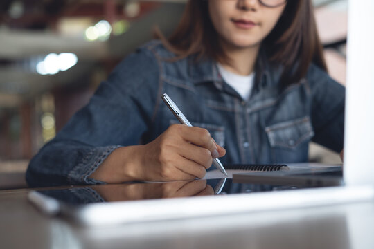 Close up of woman hand with a pen writing in notebook with laptop on desk, studying or working in a modern indoor workspace.